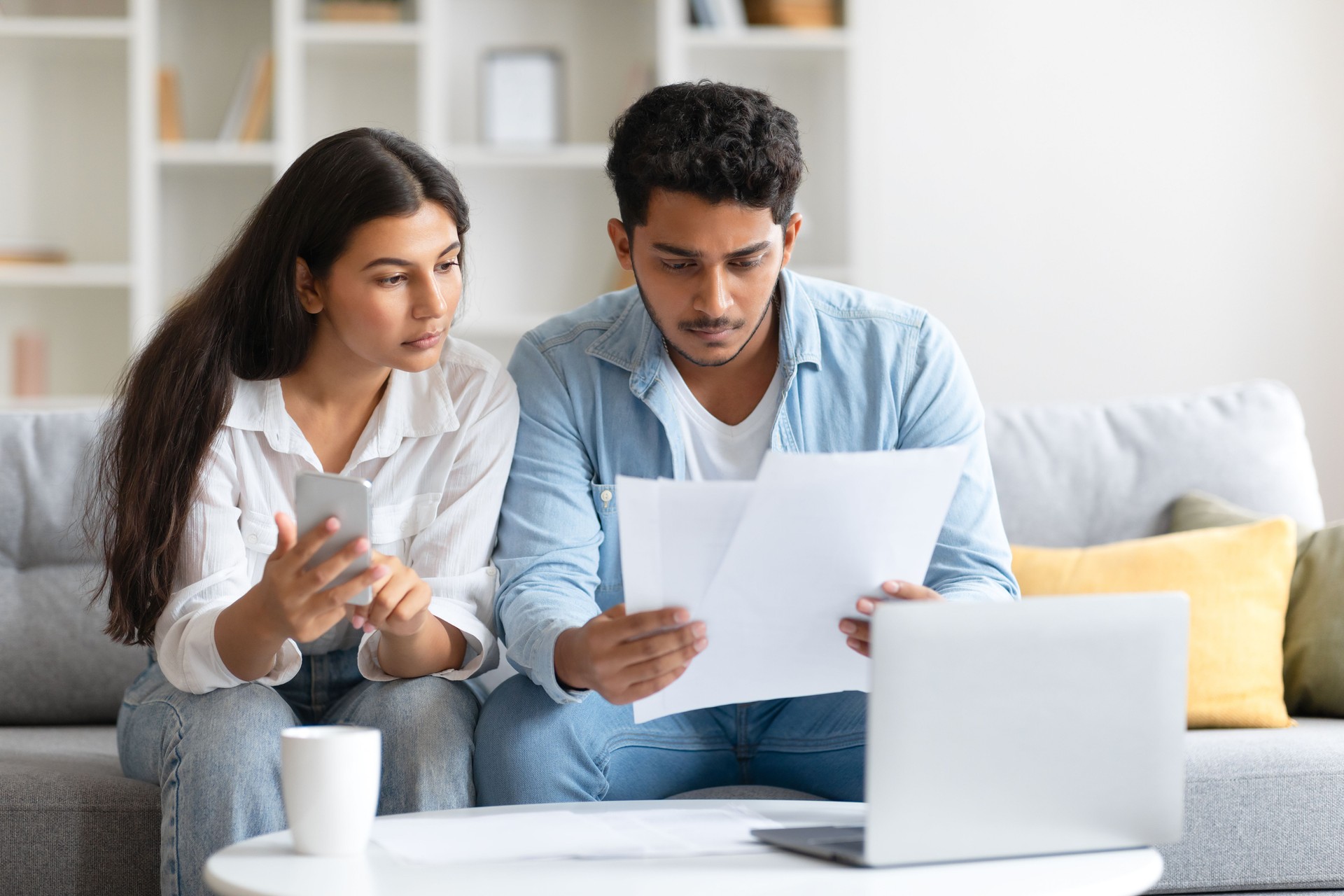 Indian couple focused on bills and using cellphone, sitting on sofa